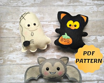 May include: Three handmade Halloween felt decorations: a white ghost with a spider, a black cat with a pumpkin, and a gray bat. The cat has orange ears and eyes. The bat has pink cheeks. The ghost has a stitched face. The image includes the text "PDF PATTERN".