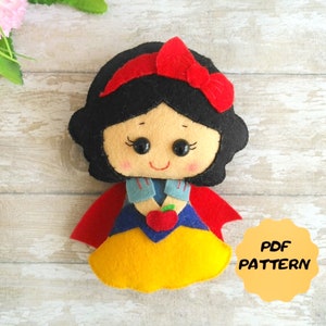 May include: Felt pattern for a Snow White doll. The doll is wearing a yellow dress, a blue shirt, a red cape, and a red bow. The doll is holding a red apple. The pattern is available as a PDF download.