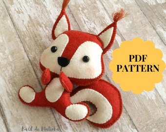 May include: A red and white felt squirrel plush toy with black eyes and a black nose. The toy is sitting on a wooden surface. The text "PDF PATTERN" is written in black on a yellow circle.