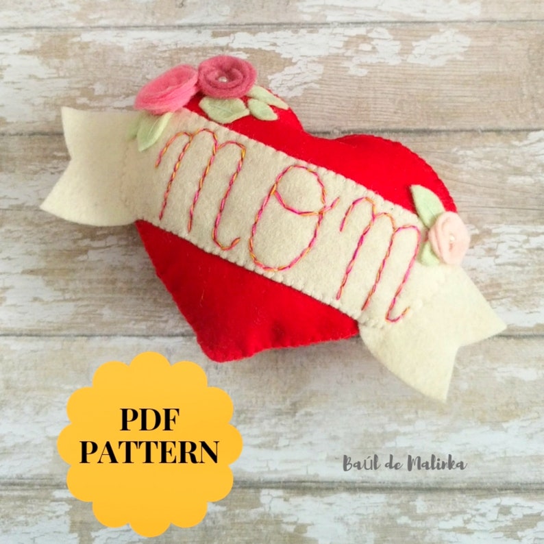 May include: A red felt heart with a white felt banner that says "mom" in pink embroidery. The banner is decorated with pink and green felt flowers. The heart is on a light wood background.