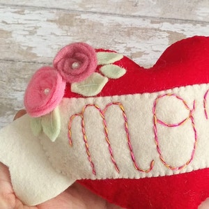May include: Red felt heart with white felt trim and the word "Mom" embroidered in pink and yellow thread. The heart is decorated with two pink felt flowers and green felt leaves.