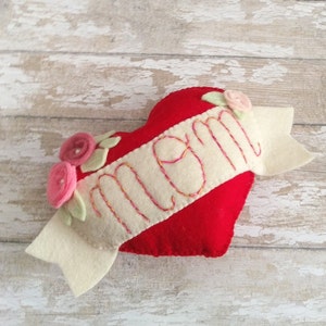 May include: A red felt heart with a white felt banner that says "Mom" in colorful embroidery. The banner has two felt flowers on the left side and one on the right.