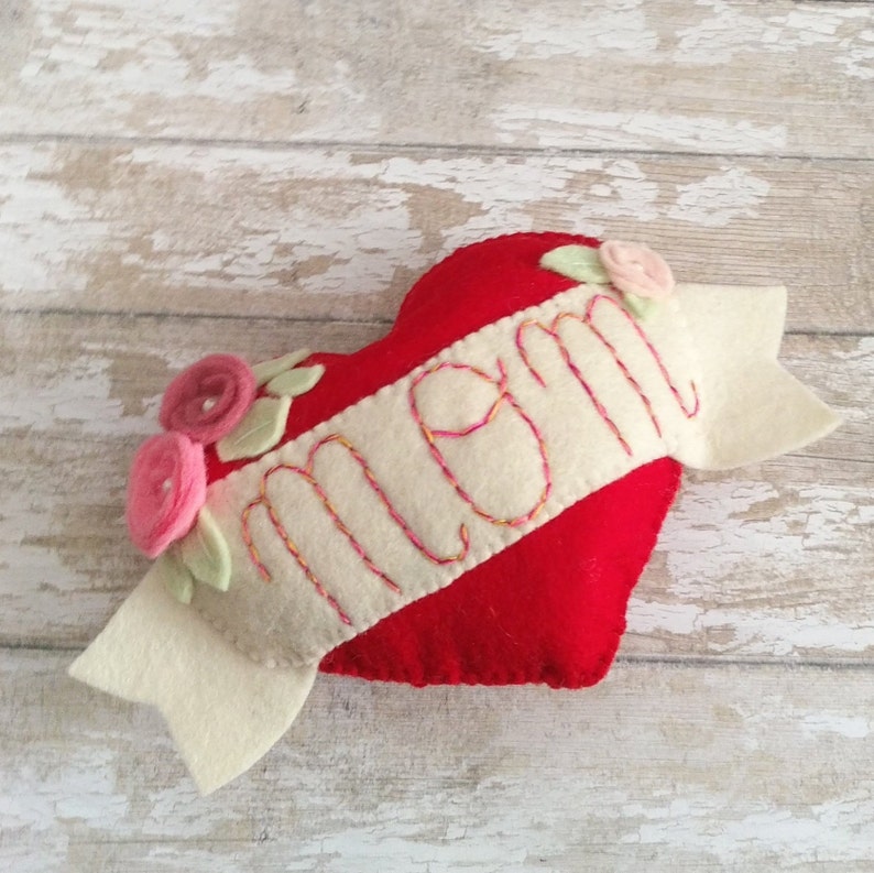 May include: A red felt heart with a white felt banner that says "Mom" in colorful embroidery. The banner has two felt flowers on the left side and one on the right.