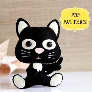 May include: A black felt cat plush toy with white accents, big eyes, and a pink nose. The toy is sitting on a white surface. The text "PDF PATTERN" is in a yellow banner above the toy.
