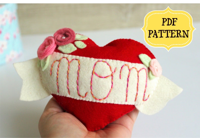 May include: A red felt heart with a white felt banner that says "MOM" in pink and orange embroidery. The heart is decorated with pink felt flowers and green felt leaves.