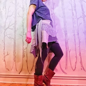 Könnte beinhalten: Eine Frau trägt einen mehrlagigen Rock in Blau und Grau mit einem abgenutzten Look. Der Rock ist in der Taille gebunden und hat einen ausgefransten Saum. Sie trägt schwarze Strumpfhosen und braune Stiefel. Der Hintergrund ist eine rosa-violette Ombre-Wand mit einem Baummuster.