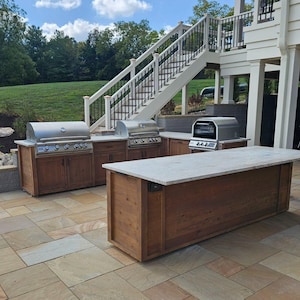 May include: Outdoor kitchen with three stainless steel grills, a large island with a white countertop, and brown wooden cabinets. The kitchen is built on a patio with tan stone tiles.