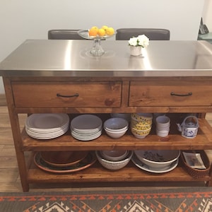 May include: A wooden kitchen island with a stainless steel countertop. The island has two drawers and two shelves. The shelves are filled with dishes and bowls. There is a small teapot on the top shelf.