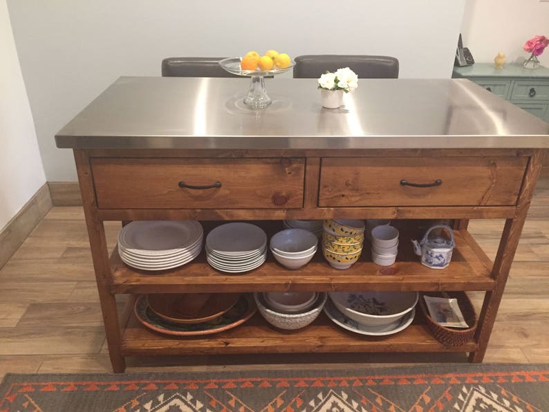 May include: A wooden kitchen island with a stainless steel countertop. The island has two drawers and two shelves. The shelves are filled with dishes and bowls. There is a small teapot on the top shelf.
