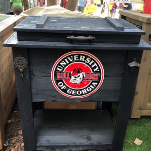 Puede incluir: Enfriador de madera negro con un estante, con un logotipo de los Bulldogs de la Universidad de Georgia. El enfriador tiene un asa de metal y un dise&ntilde;o r&uacute;stico. El enfriador est&aacute; sentado sobre una superficie de hierba verde.