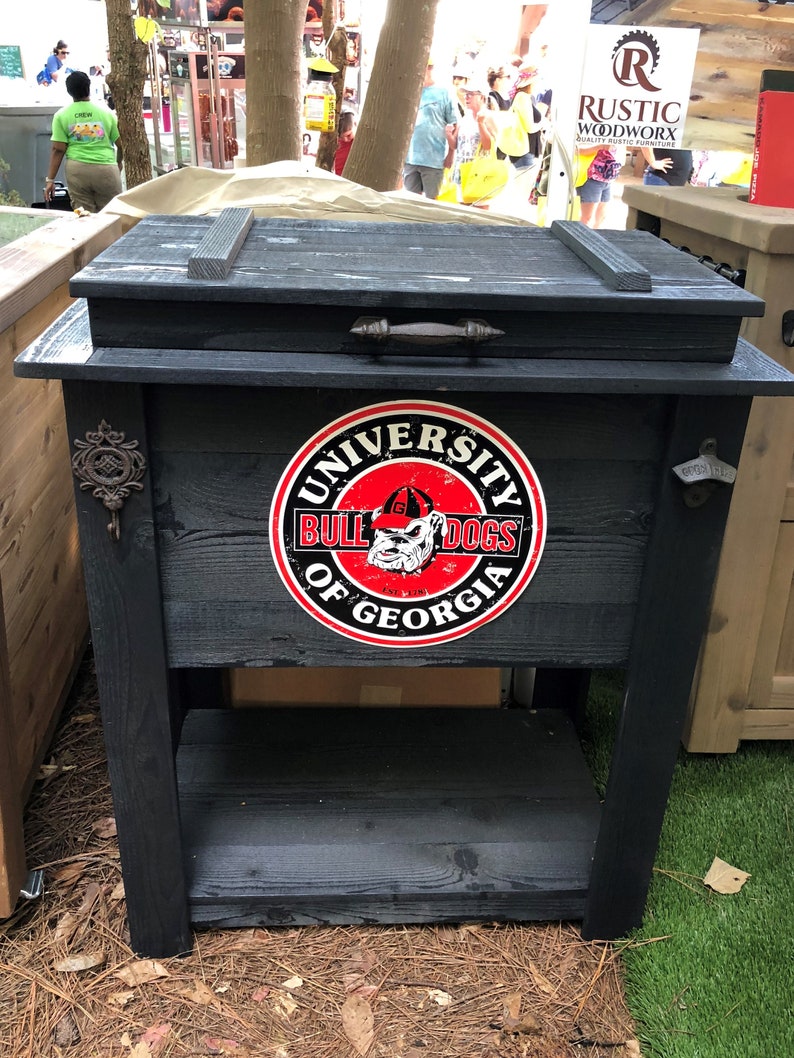 Puede incluir: Enfriador de madera negro con un estante, con un logotipo de los Bulldogs de la Universidad de Georgia. El enfriador tiene un asa de metal y un dise&ntilde;o r&uacute;stico. El enfriador est&aacute; sentado sobre una superficie de hierba verde.