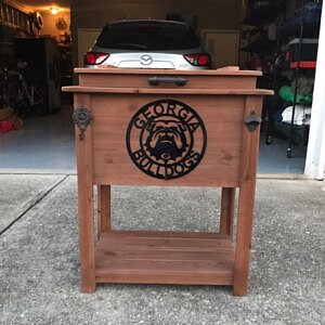 Puede incluir: Enfriador de madera con tapa de metal y un estante. El enfriador est&aacute; decorado con un recorte de metal de un bulldog con las palabras "Georgia Bulldogs" alrededor.