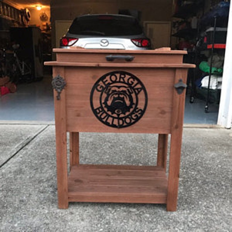Puede incluir: Enfriador de madera con tapa de metal y un estante. El enfriador est&aacute; decorado con un recorte de metal de un bulldog con las palabras "Georgia Bulldogs" alrededor.