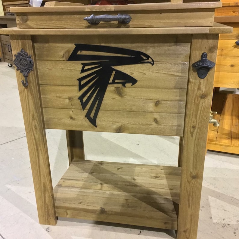 Puede incluir: Enfriador de madera con un logotipo de los Atlanta Falcons en metal negro, un abridor de botellas y un gancho. El enfriador tiene un estante debajo.