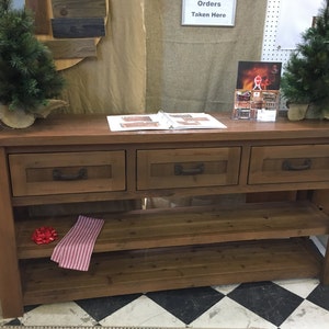 May include: A rustic wooden console table with three drawers and two shelves. The table is stained a dark brown color and has a distressed finish. The table is decorated with two small artificial pine trees.
