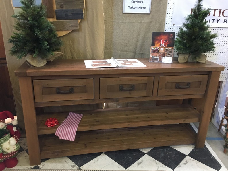 May include: A rustic wooden console table with three drawers and two shelves. The table is stained a dark brown color and has a distressed finish. The table is decorated with two small artificial pine trees.
