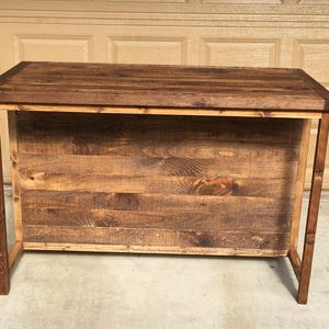 May include: A rustic wooden bar table with a dark brown stain. The table has a simple design with a rectangular top and four legs.