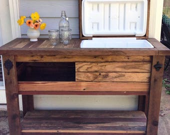 May include: A rustic wooden outdoor bar with a built-in cooler. The bar has a dark brown finish, a lower shelf, and a drawer. A bottle opener is attached to the side. A vase of yellow flowers, glasses, and a bottle are on top.