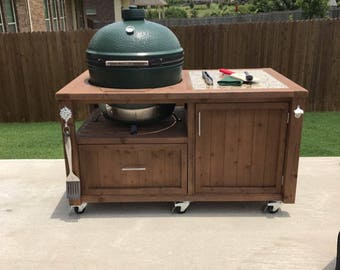 May include: A wooden outdoor grilling station with a green ceramic grill, a drawer, a cabinet with a door, and two shelves. The station has wheels and is on a concrete surface.