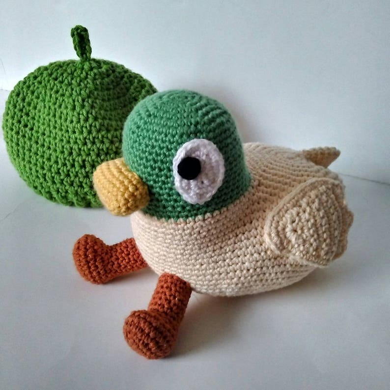 Peut inclure: Un jouet en crochet en forme de canard avec des accents verts, blancs et bruns. Le canard a un gros &oelig;il blanc avec une pupille noire. Le canard est assis sur une surface blanche.