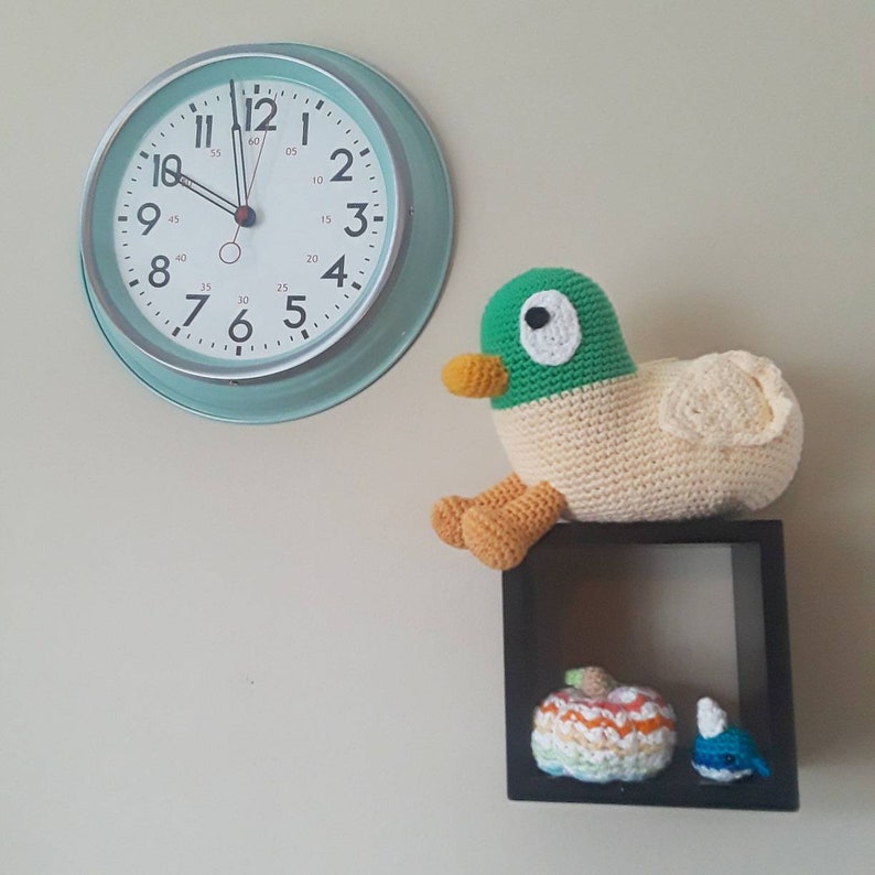 Peut inclure: Un jouet en crochet en forme de canard est plac&eacute; sur une &eacute;tag&egrave;re avec deux petites citrouilles en crochet et une baleine en crochet. Une horloge verte et blanche est accroch&eacute;e au mur derri&egrave;re l'&eacute;tag&egrave;re.