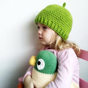 Peut inclure: Un jeune enfant portant un bonnet crochet&eacute; vert tient un jouet en peluche crochet&eacute; vert et jaune. L'enfant porte une chemise rose et est assis sur une chaise &agrave; rayures rose et blanches.