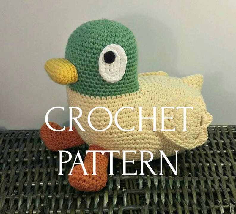 Peut inclure: Jouet en crochet amigurumi en forme de canard avec une t&ecirc;te verte, un bec jaune, un &oelig;il blanc et des pieds orange. Le jouet est pos&eacute; sur une surface en osier tress&eacute;. Le texte CROCHET PATTERN est superpos&eacute; &agrave; l'image.