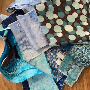 May include: A collection of blue and brown fabric scraps with various patterns, including polka dots, floral designs, and abstract patterns. The fabric scraps are arranged in a pile on a wooden surface.