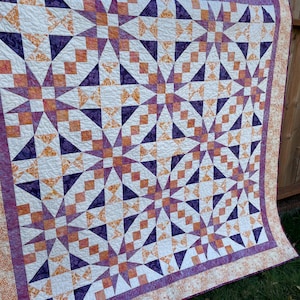 May include: A large, handmade quilt with a geometric star pattern. The quilt features white, purple, and orange fabrics. The border is a patterned orange fabric. The quilt is hanging outside on a wooden fence.