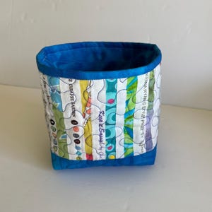 May include: A small, quilted fabric basket with a vibrant, colorful design. The exterior features vertical stripes of various patterns and colors, including green, yellow, and blue. The interior and trim are a solid bright blue. The text "Keep it Sassy" is visible.