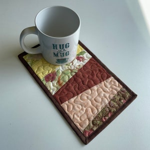 May include: A brown quilted mug rug with a white mug that says "HUG in a MUG" in teal blue lettering. The mug rug has a patchwork design with different shades of brown, beige, and green.