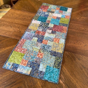 May include: A colorful patchwork table runner with a variety of geometric patterns in shades of blue, green, yellow, and red.