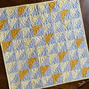 May include: A small quilt with a geometric pattern of yellow and gray triangles. The quilt is quilted with a swirling pattern.