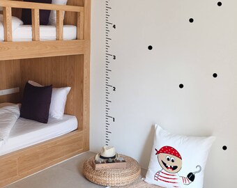 May include: A white pillow with a cartoon pirate design on it. The pillow is on a light brown rug in front of a white wall with black dots and a ruler measuring up to 6 feet.