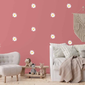 Può includere: Un muro rosa con adesivi a forma di margherita bianca, un divano letto bianco con una coperta beige e una poltrona grigia. Ci sono animali di peluche su una panca di legno di fronte al divano letto.