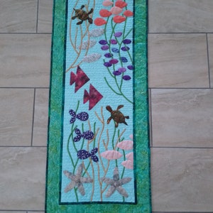 May include: A turquoise blue quilted wall hanging with a green border. The design features sea turtles, starfish, and coral in shades of pink, purple, and brown.