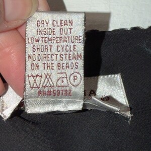 May include: A close-up of a clothing tag with care instructions. The tag is light beige with dark brown text that reads "DRY CLEAN INSIDE OUT LOW TEMPERATURE SHORT CYCLE NO DIRECT STEAM ON THE BEADS".