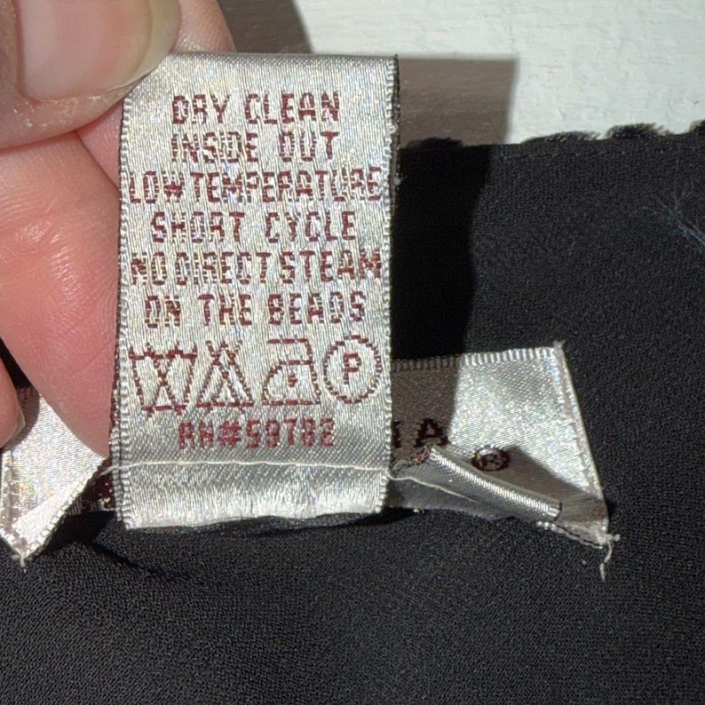 May include: A close-up of a clothing tag with care instructions. The tag is light beige with dark brown text that reads "DRY CLEAN INSIDE OUT LOW TEMPERATURE SHORT CYCLE NO DIRECT STEAM ON THE BEADS".