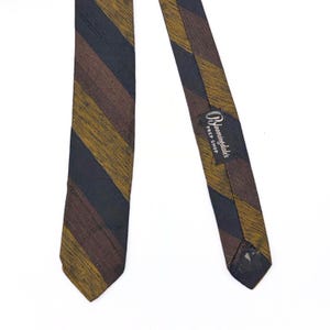 May include: A necktie featuring a diagonal striped pattern in dark navy blue, mustard yellow, and brown. The fabric has a visible textured weave. A black label on the back reads Bloomingdale's Prep Shop in white script and block lettering.