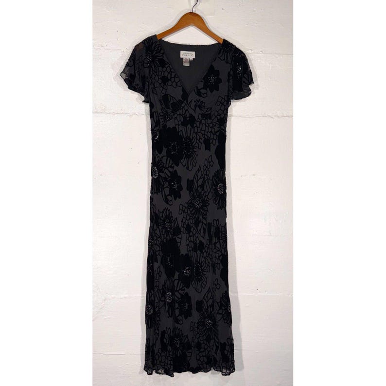 May include: A black floral print dress with a V-neck and short, flutter sleeves. The dress is floor-length and features a velvet floral pattern. The dress is hanging on a wooden hanger against a white wall.