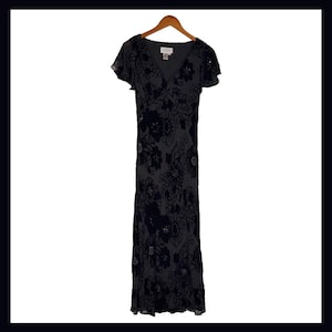 May include: A dark gray formal dress with a floral pattern. The dress has a V-neck, short flutter sleeves, and a floor-length skirt. The floral design is in a darker shade, creating a subtle contrast. The dress is hanging on a wooden hanger.