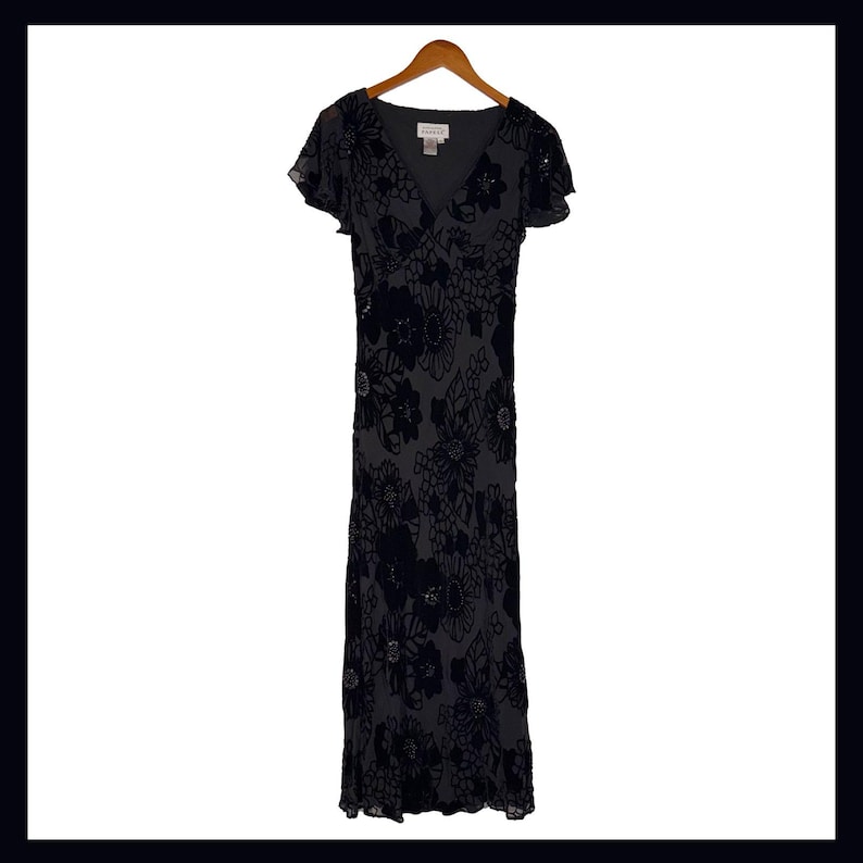 May include: A dark gray formal dress with a floral pattern. The dress has a V-neck, short flutter sleeves, and a floor-length skirt. The floral design is in a darker shade, creating a subtle contrast. The dress is hanging on a wooden hanger.