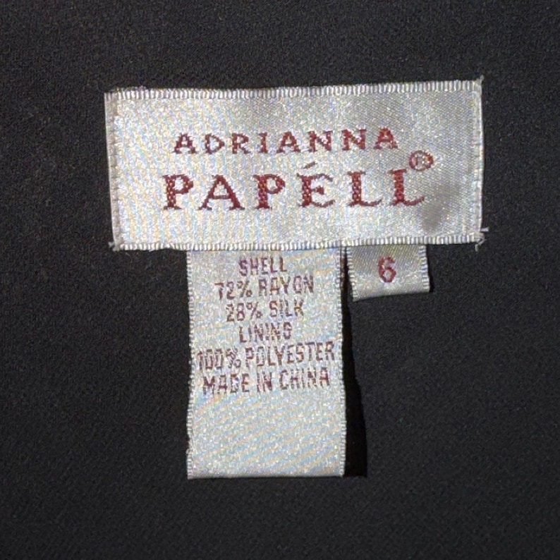 May include: A close-up of a clothing tag from the brand Adrianna Papell. The tag is rectangular with the brand name in a serif font. The tag lists the shell as 72% rayon and 28% silk, with a 100% polyester lining. The size is 6.