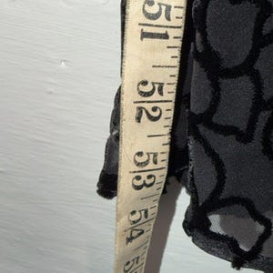 May include: A black garment with a floral pattern is measured with a vintage measuring tape. The tape shows measurements from 51 to 54 inches. The fabric has a velvet-like texture.