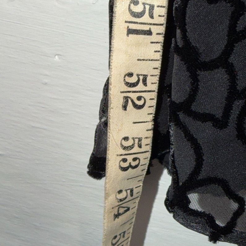 May include: A black garment with a floral pattern is measured with a vintage measuring tape. The tape shows measurements from 51 to 54 inches. The fabric has a velvet-like texture.