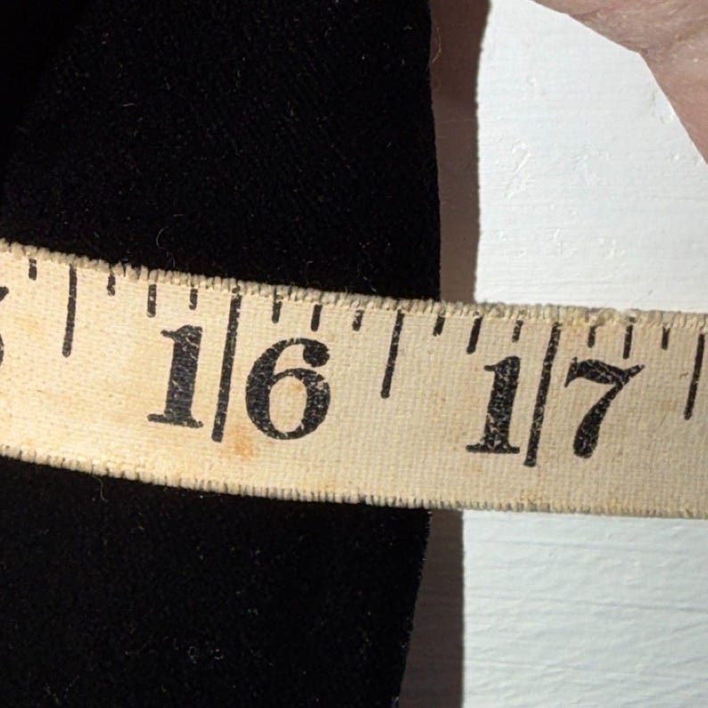 May include: A beige measuring tape showing the number 16 and 17 inches. The tape is placed next to a black fabric. The tape is slightly worn and has black markings.