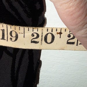 May include: A black fabric is measured with a vintage measuring tape. The tape shows a measurement of 20 inches. The fabric appears to be textured and possibly velvet.