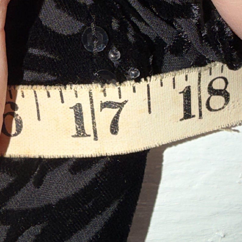 May include: Close-up of black fabric with a textured pattern and sequins. A beige measuring tape shows the measurement between 17 and 18 inches.