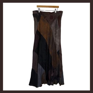 May include: A long, ankle-length skirt featuring a patchwork design with various panels in shades of brown, black, and grey. The skirt is constructed from multiple fabric sections with different textures and patterns, hanging from a wooden hanger.