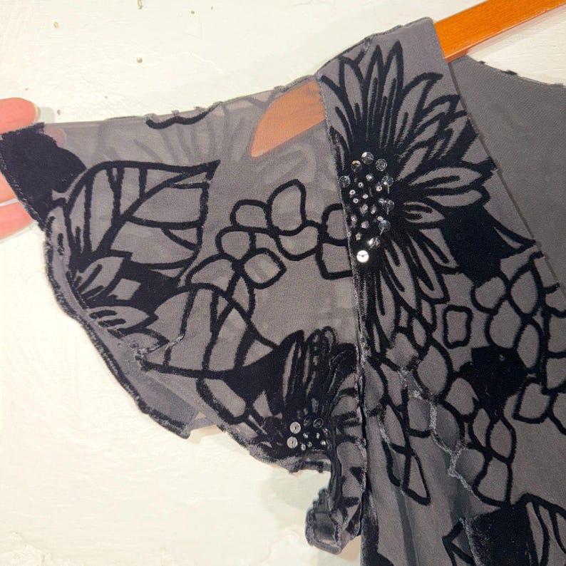 May include: Close-up of a gray garment with black floral velvet patterns. The sheer fabric reveals a glimpse of the arm and shoulder. The neckline is embellished with sequins.